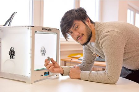 Ein lächelnder junger Mann hält eine 3D-gedruckte Figur neben einem Ultimaker 3D-Drucker.