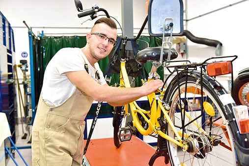 Ein junger Handwerker arbeitet in einer Zweiradwerkstatt an einem gelben Fahrrad.