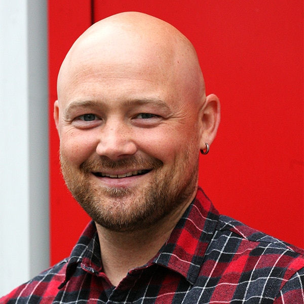 Porträt von Jan Langer im rot-schwarz karierten Hemd vor rotem Hintergrund.