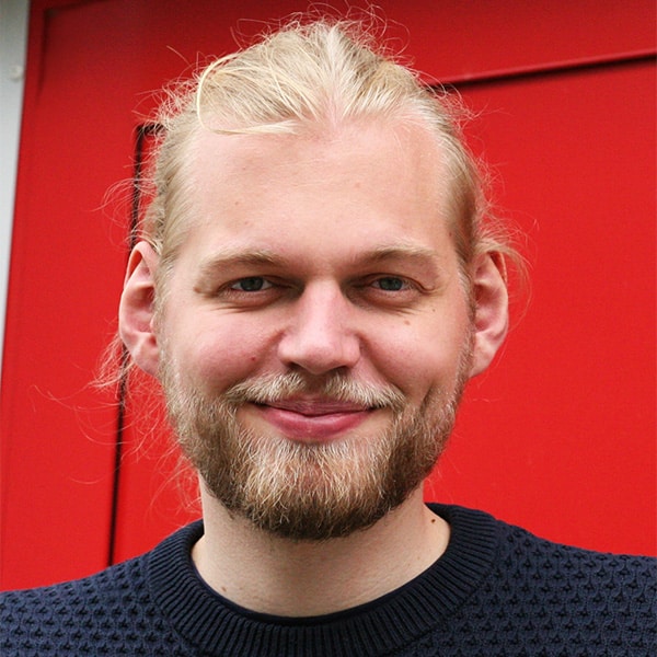 Porträt von Lukas Schütt mit Bart und blondem Haar vor einem roten Hintergrund.