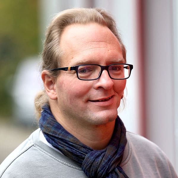 Porträt von Carsten mit Brille, blauem Schal und zurückgebundenem Haar.