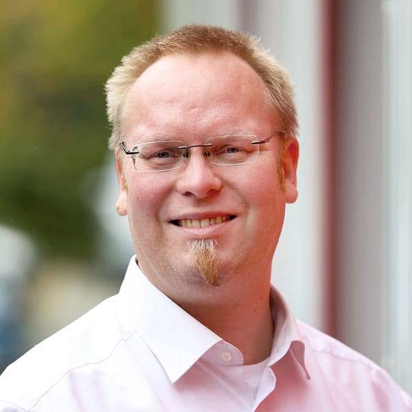 Porträt von Sven Niebelshütz mit Brille und kurzem Bart in einem weißen Hemd.